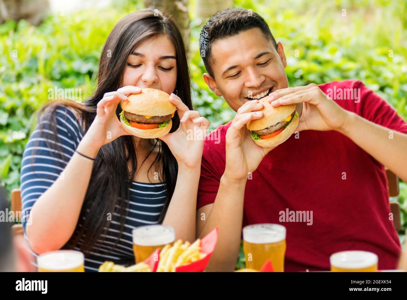 Couple heureux de manger des hamburgers et de boire de la bière glacée froide dans un restaurant en plein air dans une vue rapprochée d'eux souriant en anticipation de leur morsure Banque D'Images