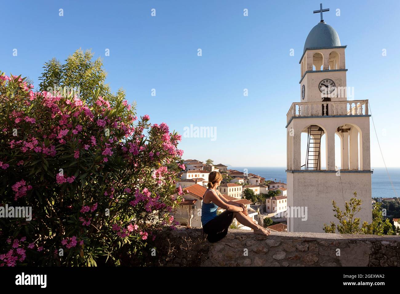 Femme assise près d'une oléande fleurie sur un petit mur, regardant le clocher de l'église et belle vue sur la mer et le village, Dhermi, Albanie Banque D'Images