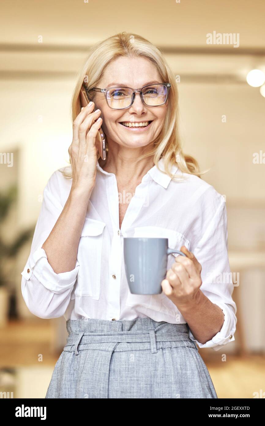 Portrait d'une femme d'affaires blonde mûre et attirante souriant à l'appareil photo tout en faisant un appel et en buvant un café, debout dans son bureau Banque D'Images
