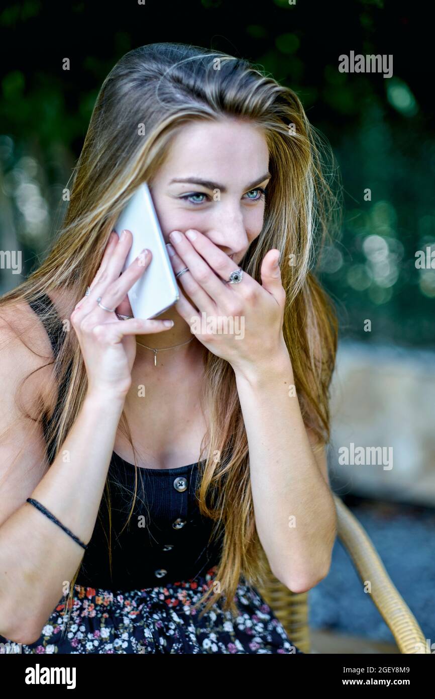 Portrait de beauté naturelle d'une jeune femme dans ses années 20 avec de longs cheveux et des yeux bleus à l'extérieur dans un jardin parlant sur son téléphone mobile. Concept de style de vie. Banque D'Images