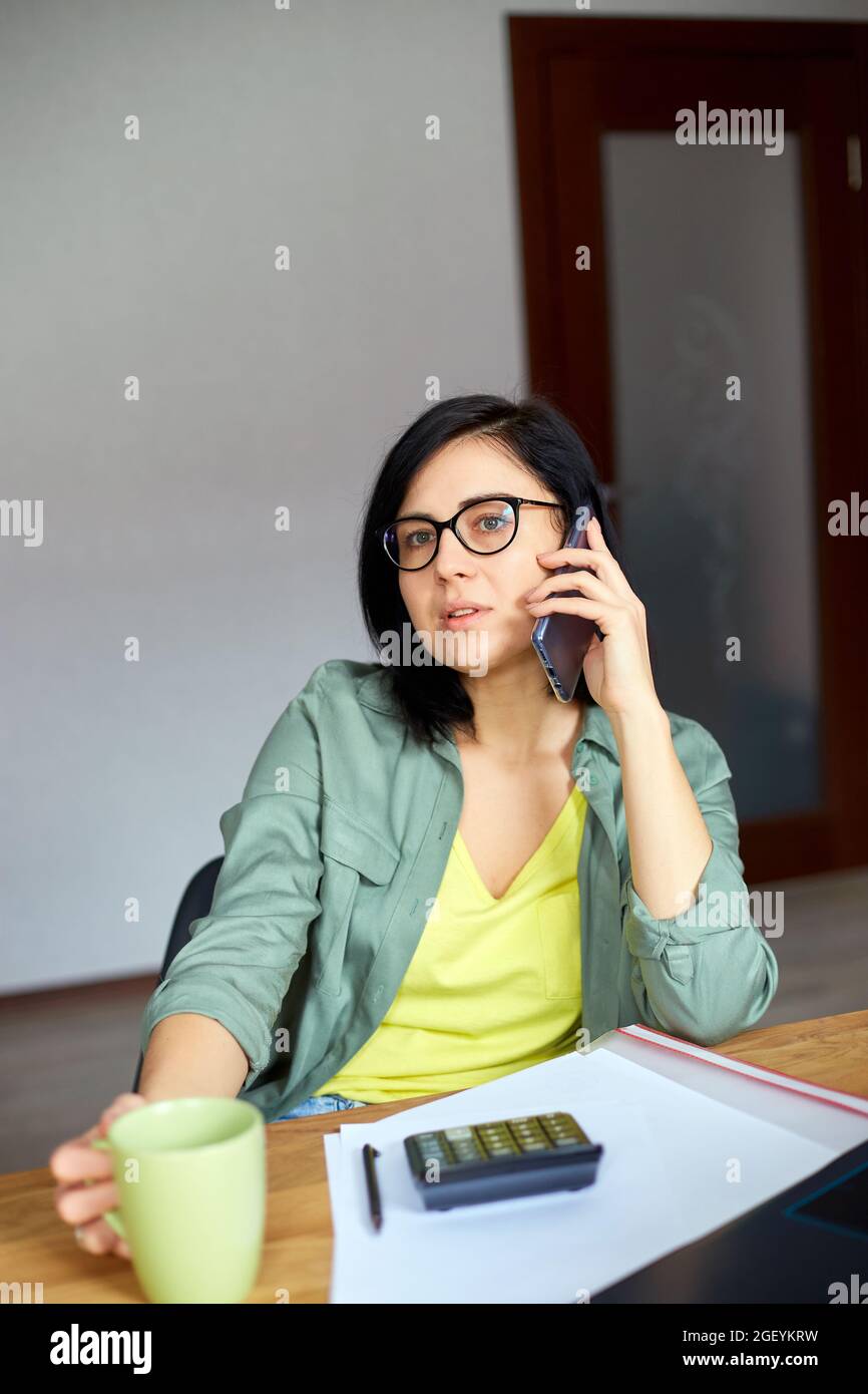 Femme brune élégante en lunettes assise à une table en bois avec bloc-notes  et ayant un appel téléphonique, boire du thé dans un lieu de travail  moderne, indépendant travaillant à hom Photo