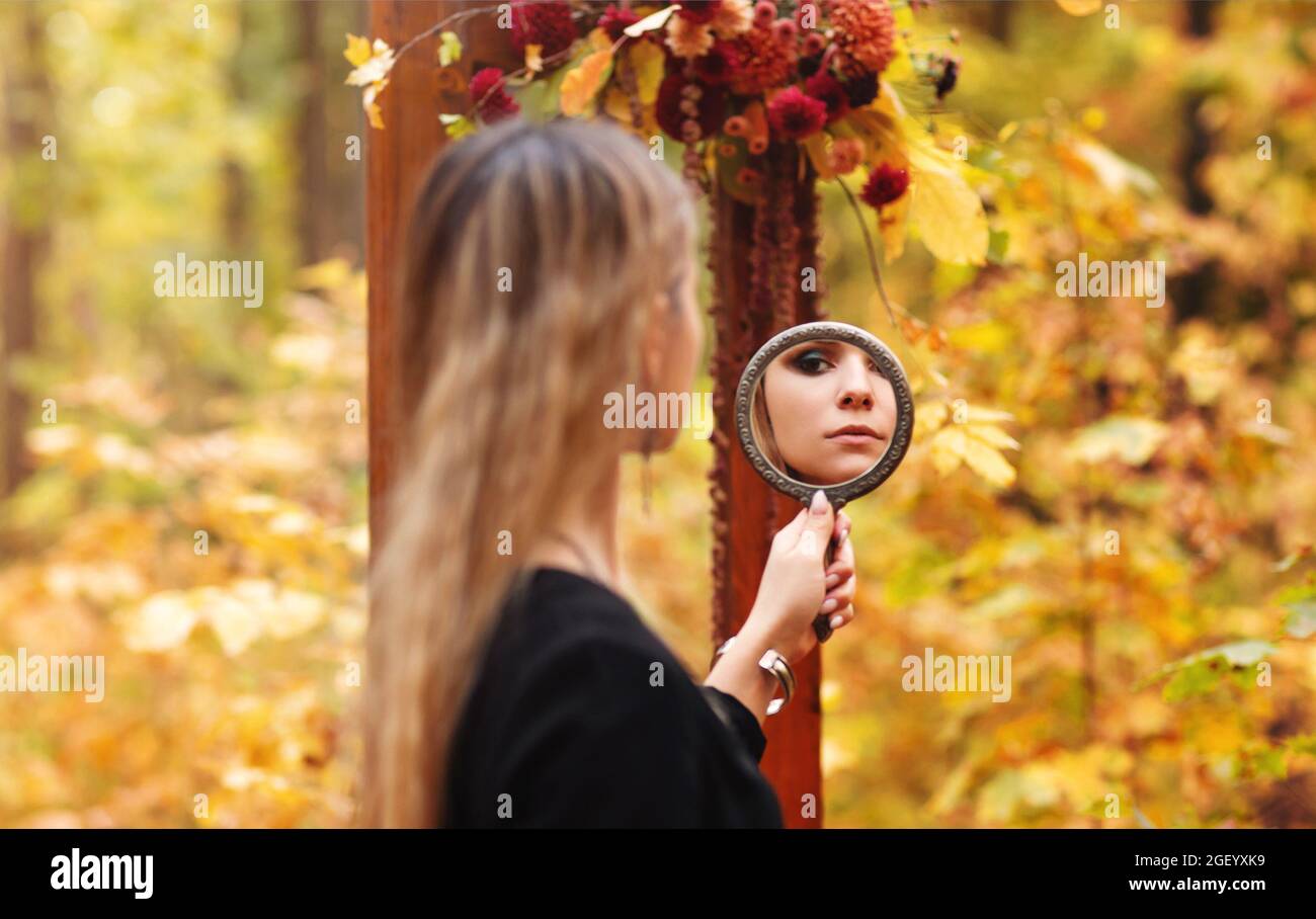 Photo pittoresque de la jeune fille belle avec maquillage du soir portant le costume de sorcière noir tenant le miroir et le regardant pendant la séance photo d'halloween i Banque D'Images