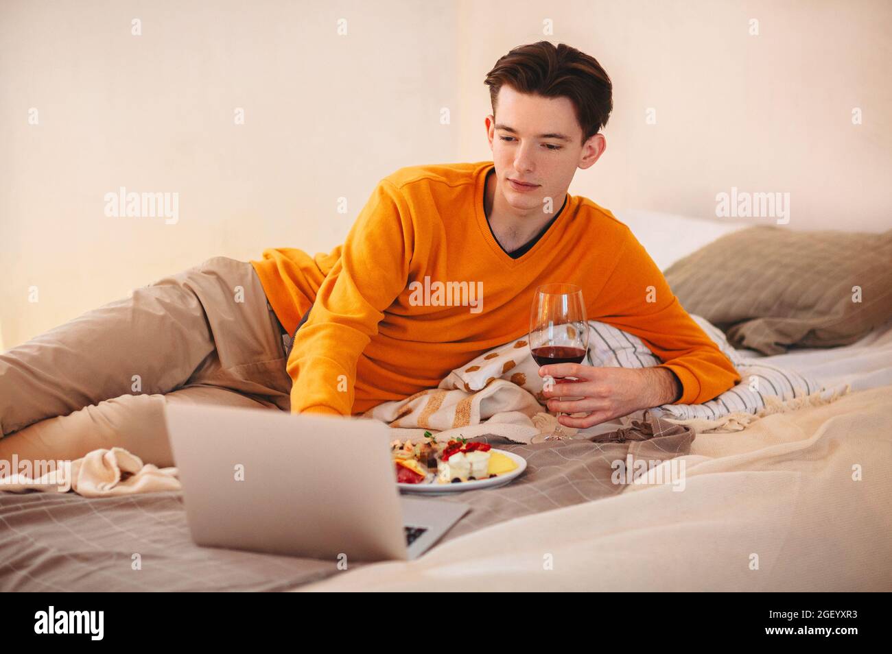 Jeune homme décontracté avec un verre de vin dans les mains, allongé sur un lit confortable avec une assiette de nourriture, regardant des vidéos sur un ordinateur portable tout en mangeant, en appréciant le leais Banque D'Images