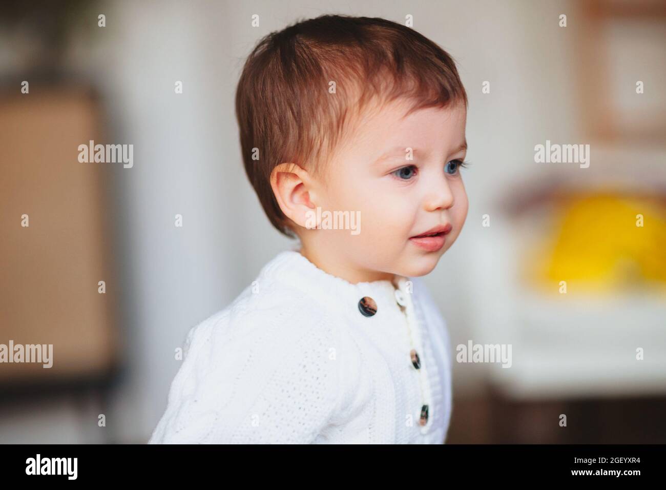 Portrait d'un adorable petit garçon de rêve avec des yeux bleus portant un chandail blanc à boutons sur les côtés en se tenant à l'intérieur, à l'intérieur Banque D'Images