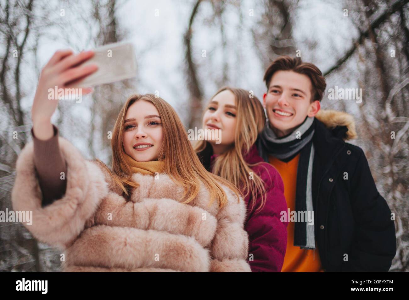 Image de trois jeunes heureux heureux meilleurs amis, deux filles et un garçon faisant selfie sur téléphone mobile en souriant largement sur l'appareil photo, debout contre neige-cov Banque D'Images