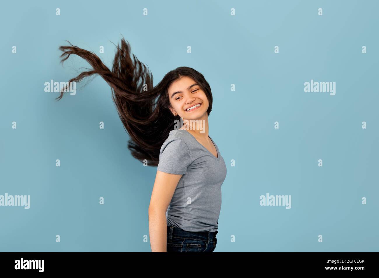 Portrait d'une adolescente indienne souriante posant avec de longs cheveux de vol sombres sur fond bleu studio Banque D'Images