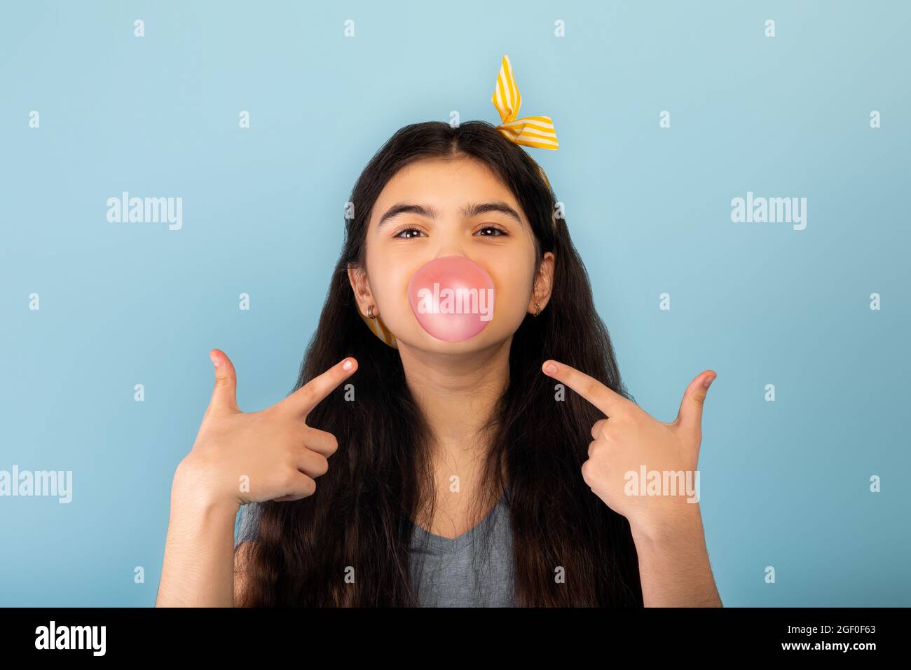 Drôle indienne jeune fille soufflant de la bulle de sucre sans chewing-gum, pointant vers elle sur fond bleu de studio Banque D'Images