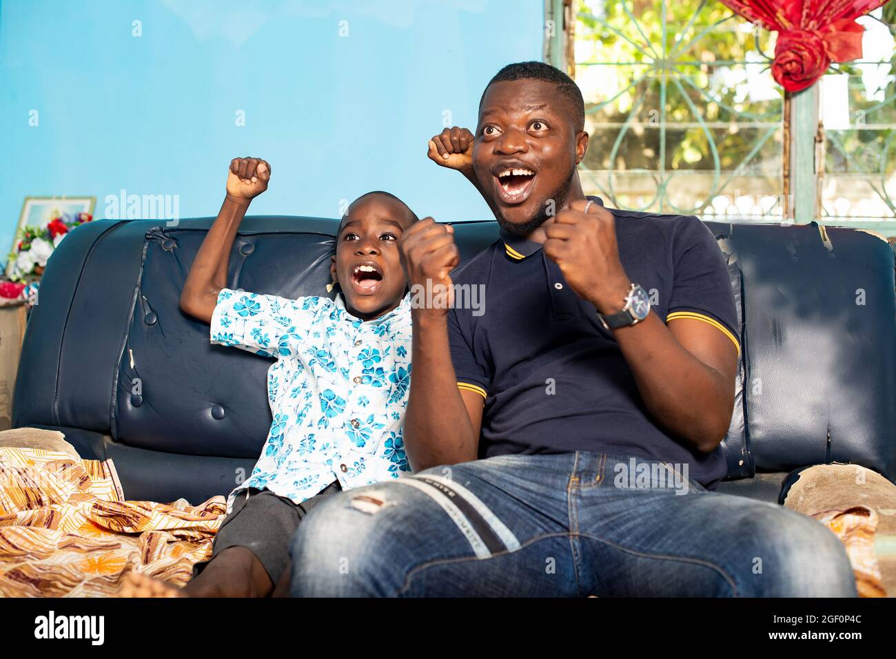 Le jeune père africain portant un polo bleu et un Jean et son fils d'âge  préscolaire vêtu d'une belle chemise bleue sont assis dans le même fauteuil  à l'intérieur du h Photo
