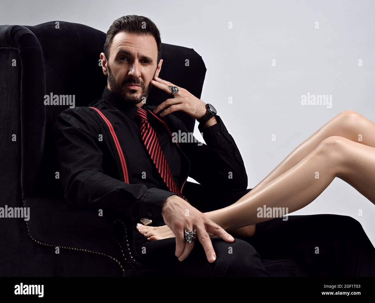 Arrogant homme brutal en chemise noire avec cravate et bretelles assis dans  un grand fauteuil avec des jambes de femme couché sur le genou Photo Stock  - Alamy