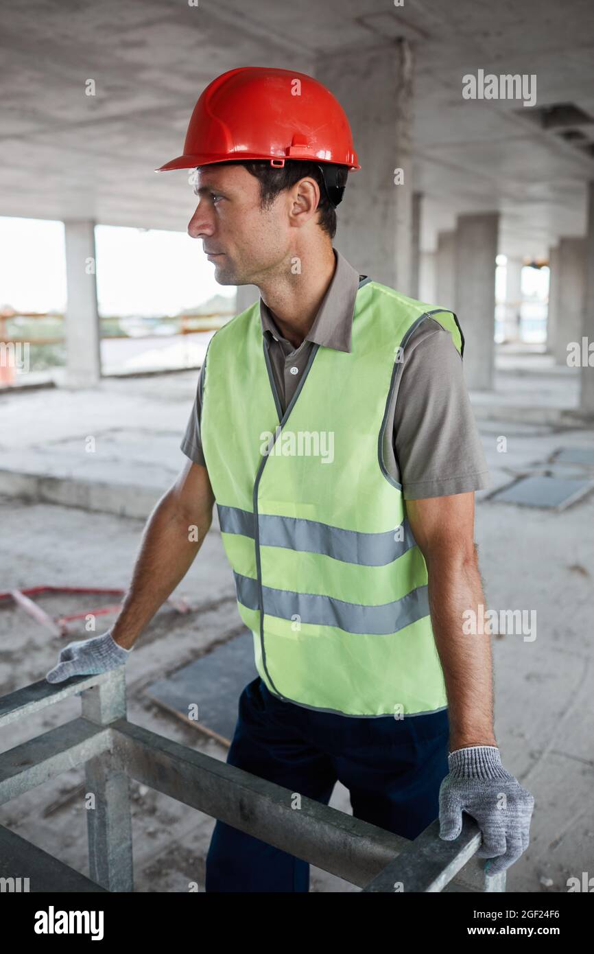 Portrait vertical de l'ouvrier de construction masculin portant un casque et une veste réfléchissante sur le chantier et regardant loin Banque D'Images