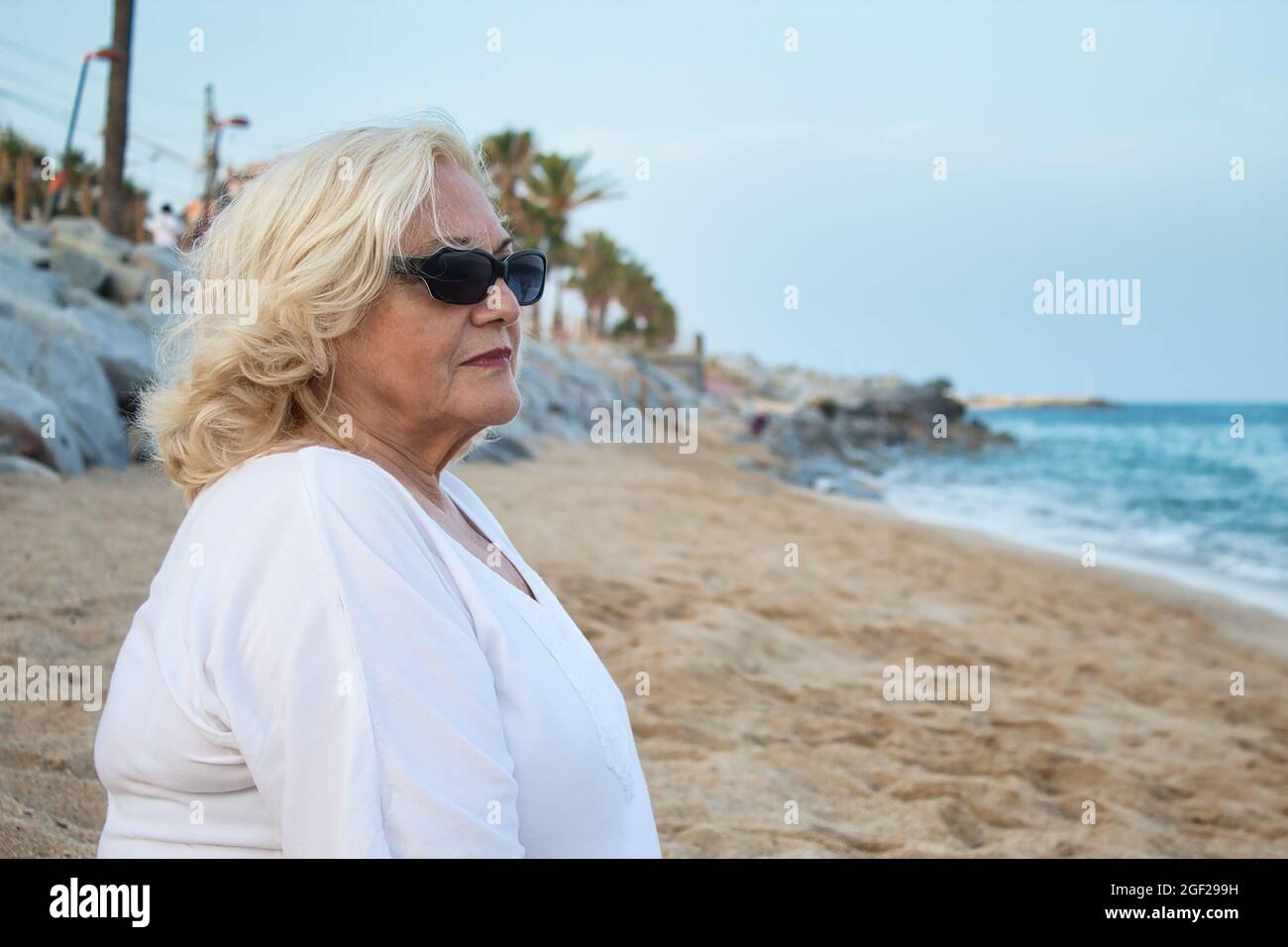 femme mature en lunettes de soleil assise seule sur la plage au bord de la mer dans une robe blanche Banque D'Images