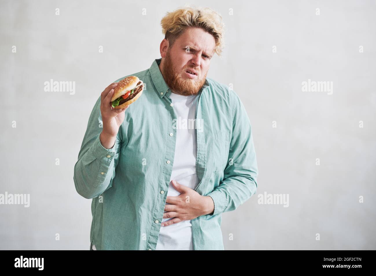 Homme en surpoids ayant une douleur à l'estomac de manger de la nourriture rapide contre le fond blanc Banque D'Images
