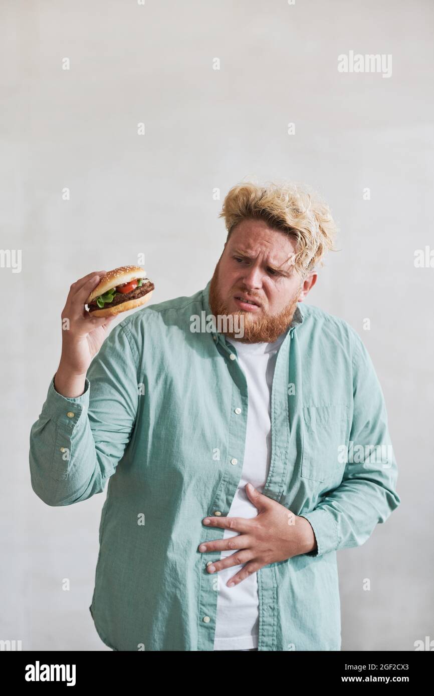Un jeune homme en surpoids regardant un hamburger dans sa main et ayant mal à l'estomac de la restauration rapide isolée sur fond blanc Banque D'Images