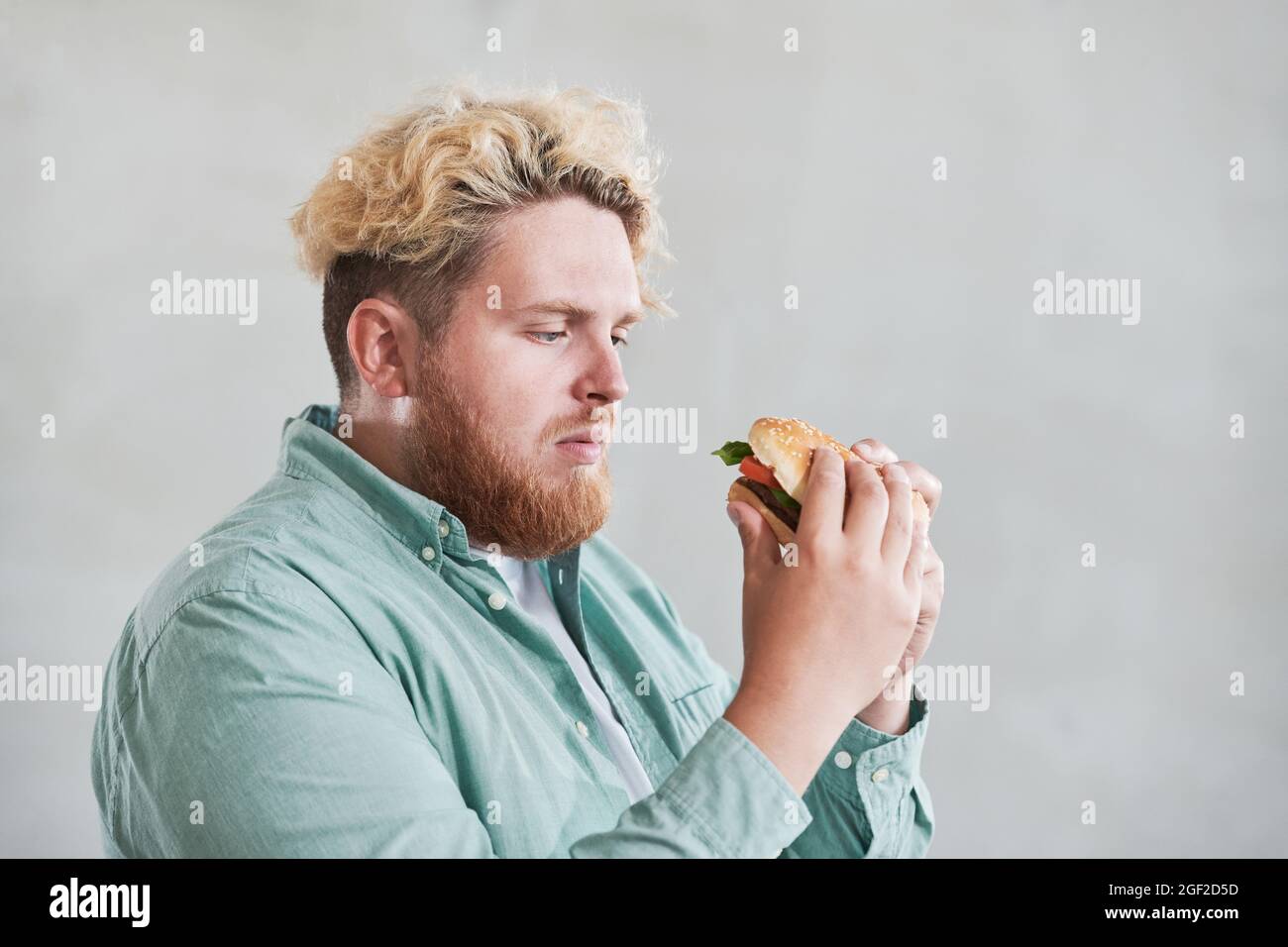 Homme en surpoids regardant le hamburger dans ses mains avec triste contre le fond blanc qu'il s'est assis sur un régime Banque D'Images