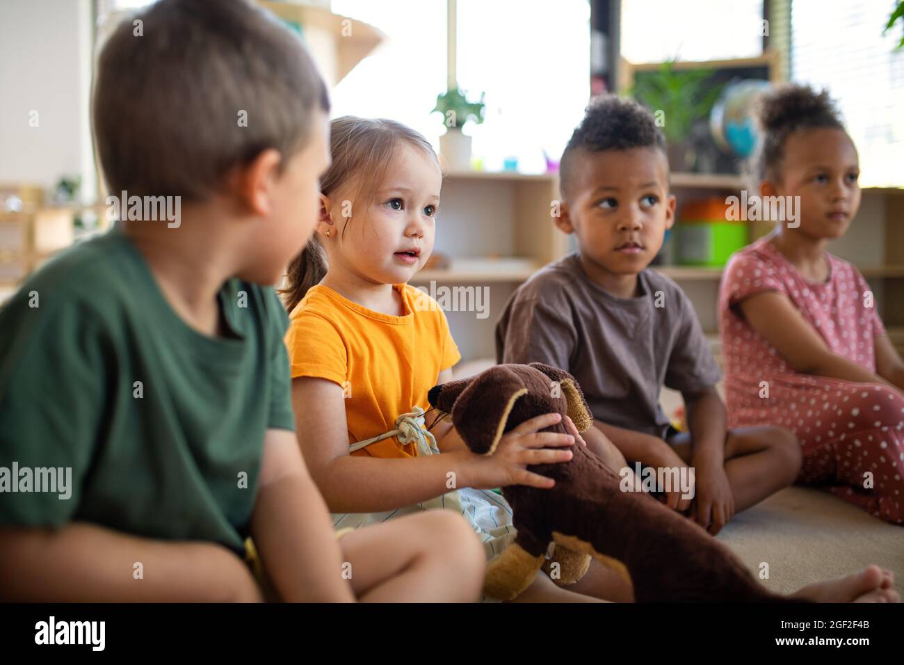 Groupe de petits enfants de la maternelle assis sur le sol à l'intérieur dans la salle de classe, à l'écoute de l'enseignant. Banque D'Images