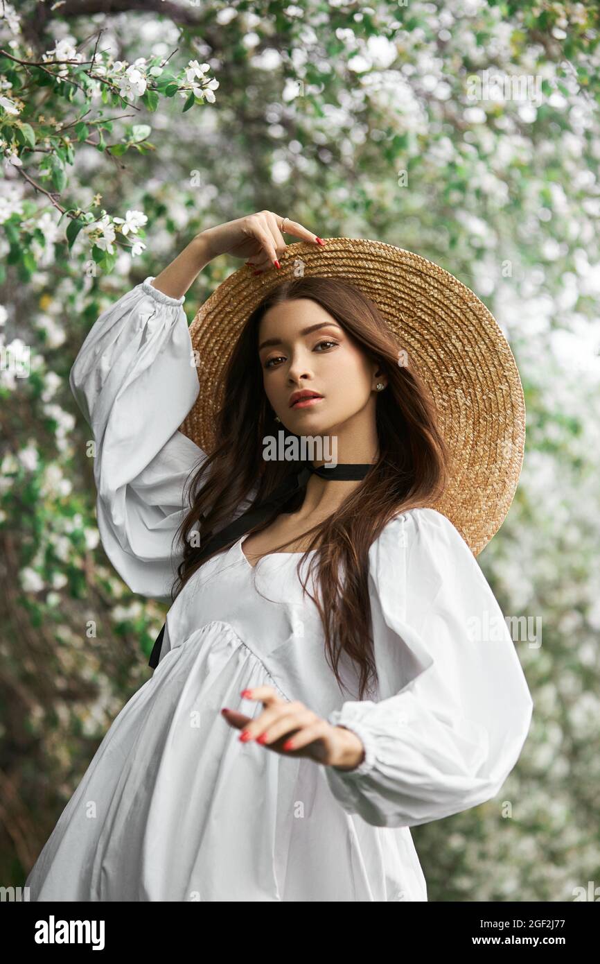 Femme aux cheveux bruns dans un grand chapeau de paille et une robe blanche  pose sur le fond des arbres blancs en fleurs. Look romantique, beauté  naturelle, propre f Photo Stock -