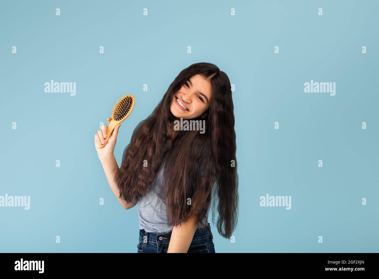 Jolie jeune fille indienne avec des cheveux longs soyeux foncés tenant une brosse en bois, posant sur fond bleu studio Banque D'Images