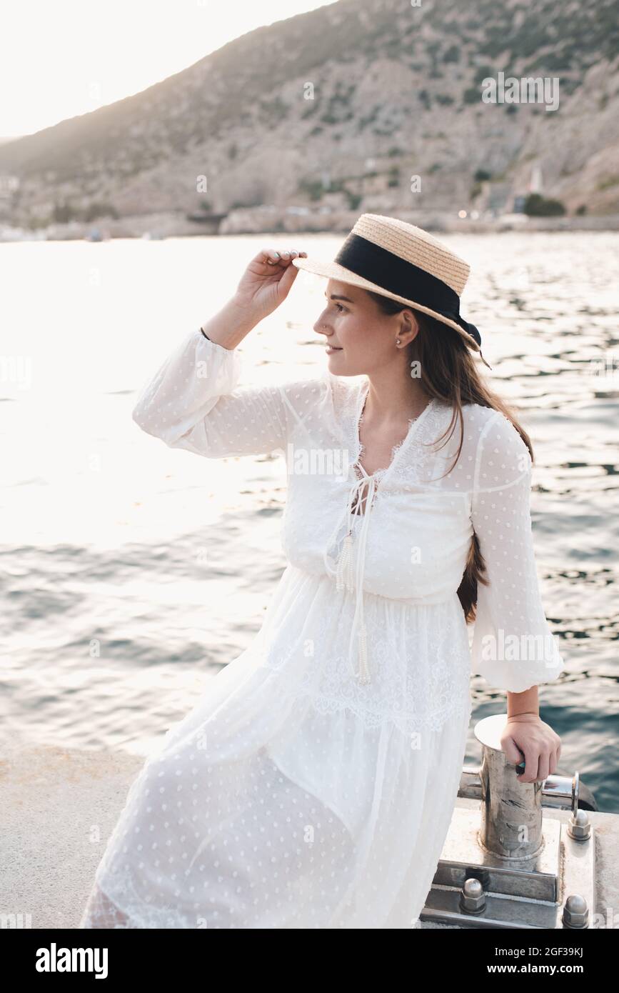 Jeune femme élégante 24-26 ans porter chapeau de paille et blanc élégant  robe posant au-dessus de la mer à quai en plein air. Saison des vacances d' été. Femme romantique Photo Stock -