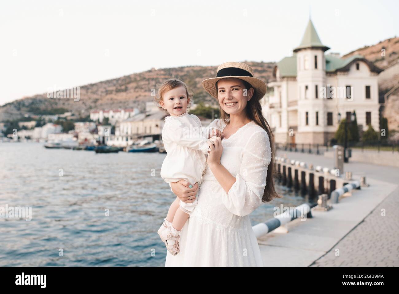 Joyeux Bebe Fille Gai Assis Sur Les Mains De La Mere Porter Un Elegant Blanc Dresse Et Chapeau De Paille Se Detendre Sur La Cote De Mer A Quai Dehors De Pres