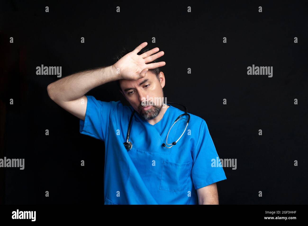 Médecin vêtu d'un uniforme bleu d'hôpital, sombre et barbu, portant un stéthoscope autour de son cou, essuyant son front avec sa main avec un ou fatigué Banque D'Images