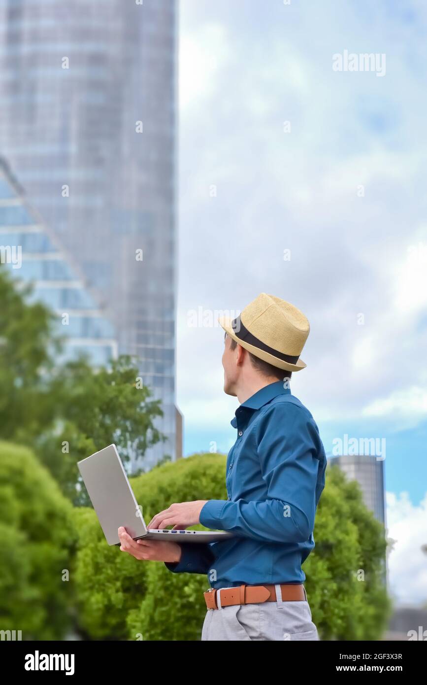 Un jeune homme avec un ordinateur portable travaille à distance à l'extérieur et regarde vers le bâtiment de bureau. Travailler et étudier à distance en raison de restrictions en cas de pandémie. Banque D'Images