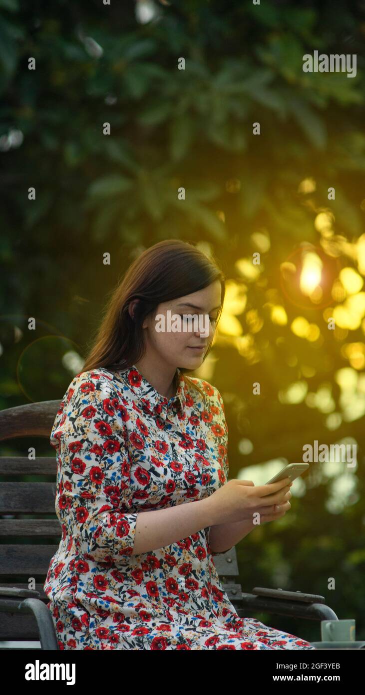 Belle jeune femme latine en robe blanche à motif floral lisant et tapant sur son smartphone dans le jardin au coucher du soleil avec les rayons du soleil passant thro Banque D'Images
