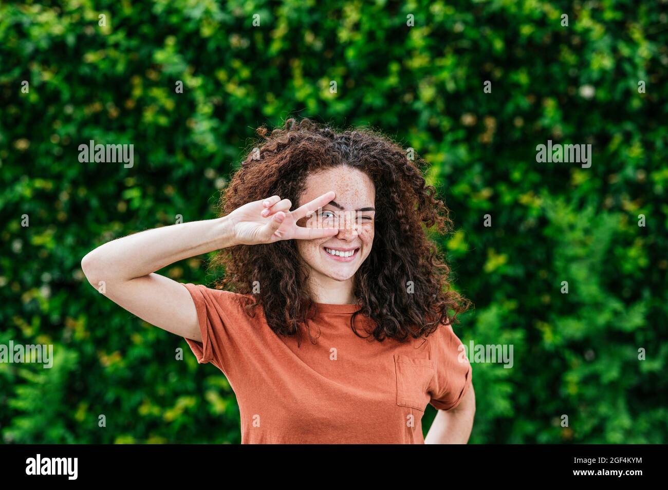 Une jeune femme souriante se tord tout en faisant signe de paix devant des plantes de lierre vertes Banque D'Images