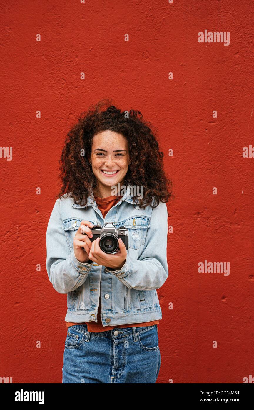 Femme souriante tenant l'appareil photo devant le mur rouge Banque D'Images