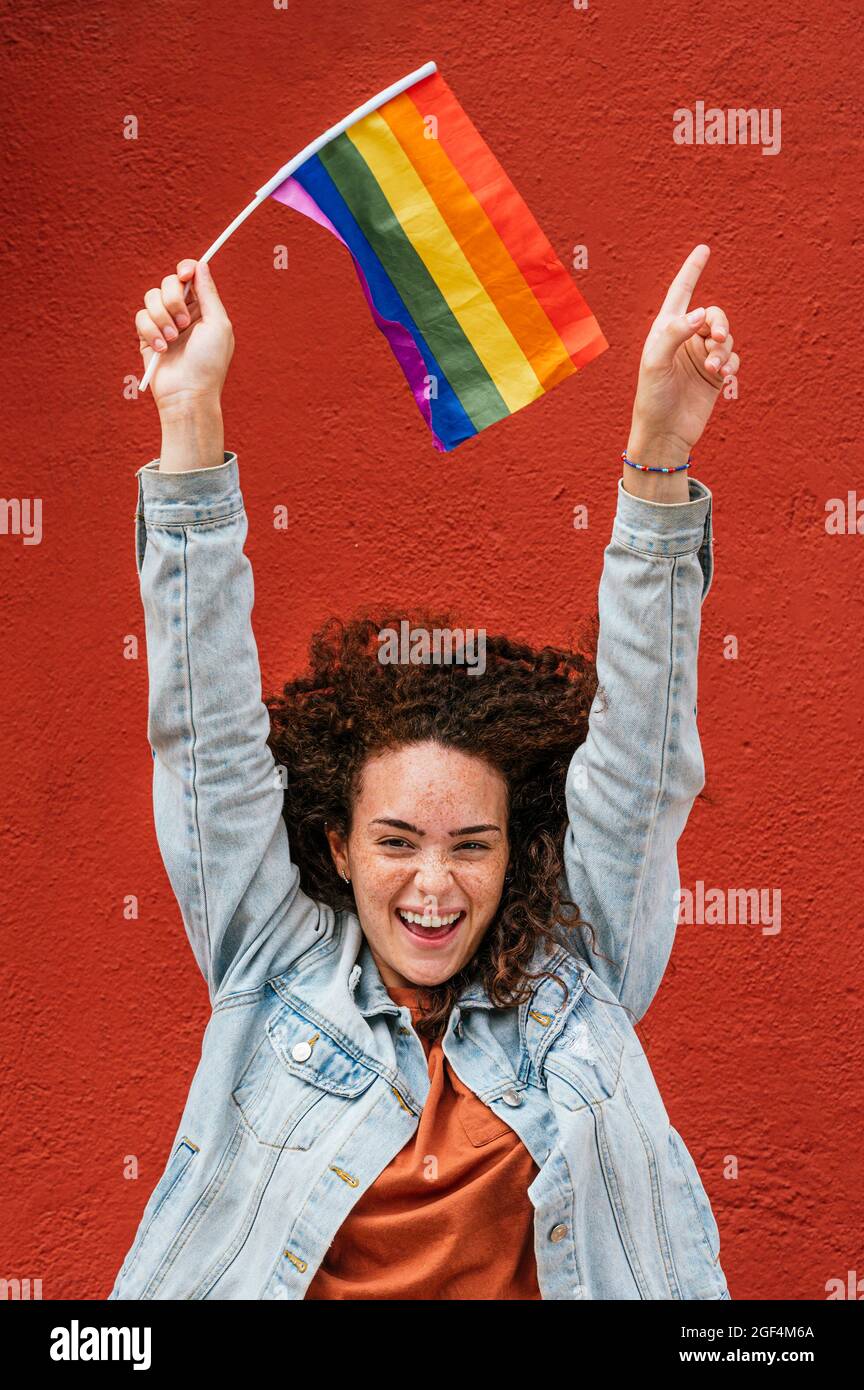 Bonne femme aux cheveux bouclés avec drapeau arc-en-ciel devant le mur rouge Banque D'Images