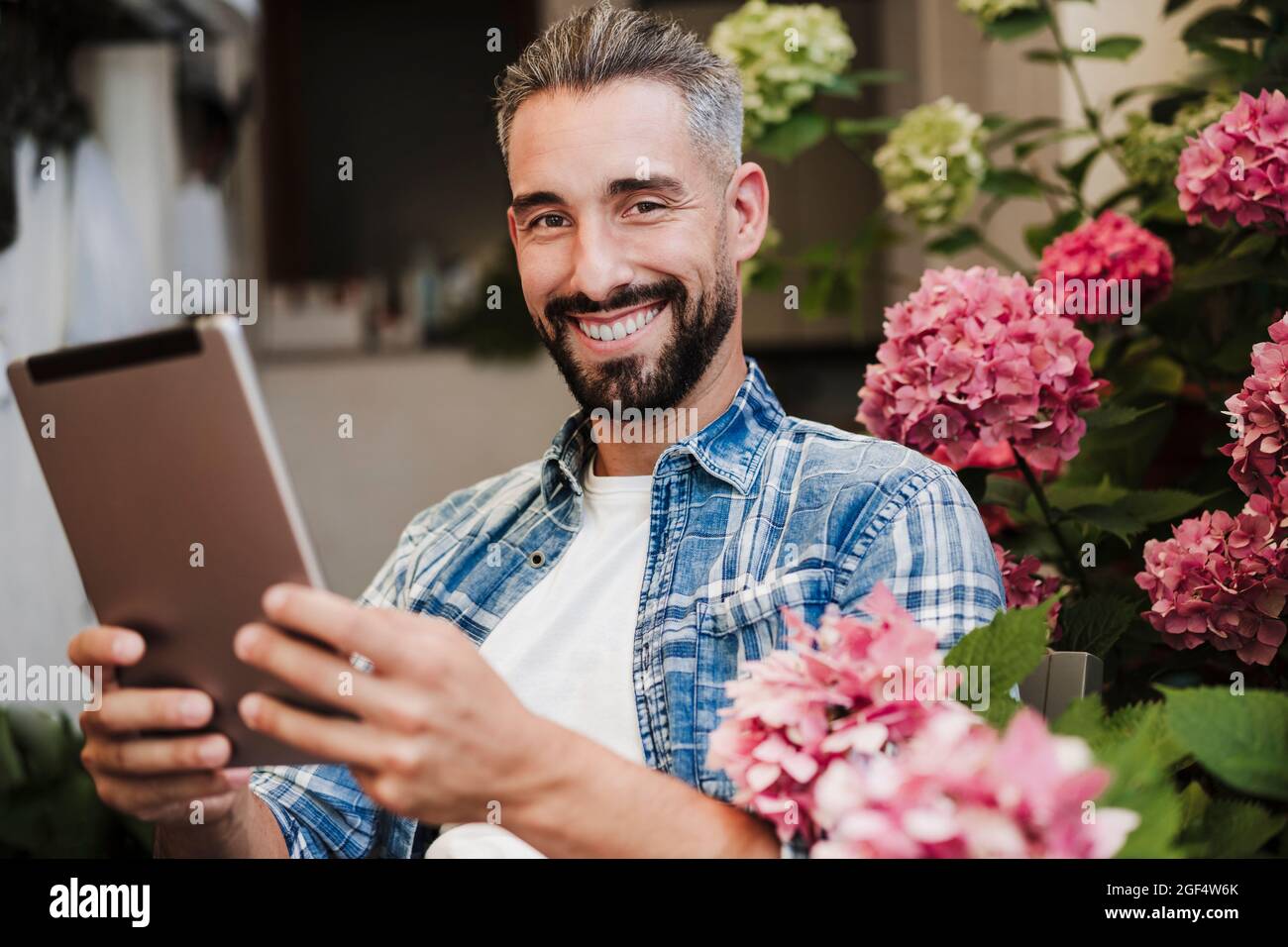 Beau travailleur indépendant masculin tenant une tablette numérique par une plante à fleurs dans l'arrière-cour Banque D'Images