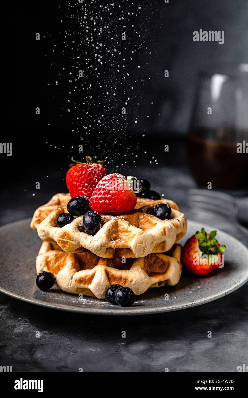 Sucre en poudre tombant sur une assiette de gaufres fraîches avec fraises et bleuets Banque D'Images