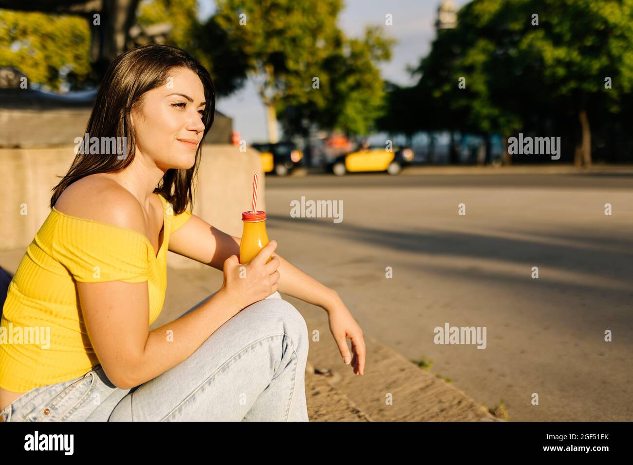Jeune femme avec une bouteille de jus de fruit regardant loin tout en étant assise sur le sentier Banque D'Images