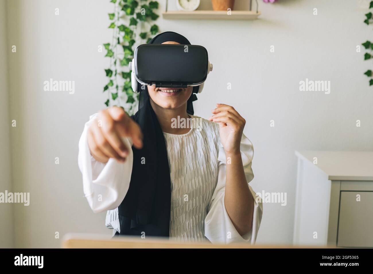 Souriante employée freelance pointant tout en utilisant un casque de réalité virtuelle à la maison Banque D'Images