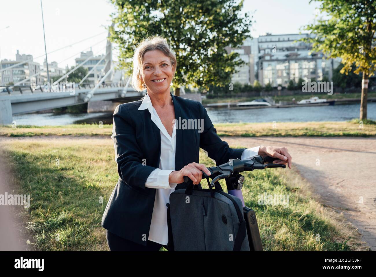 Femme souriante professionnelle avec scooter électrique poussant debout sur l'herbe Banque D'Images