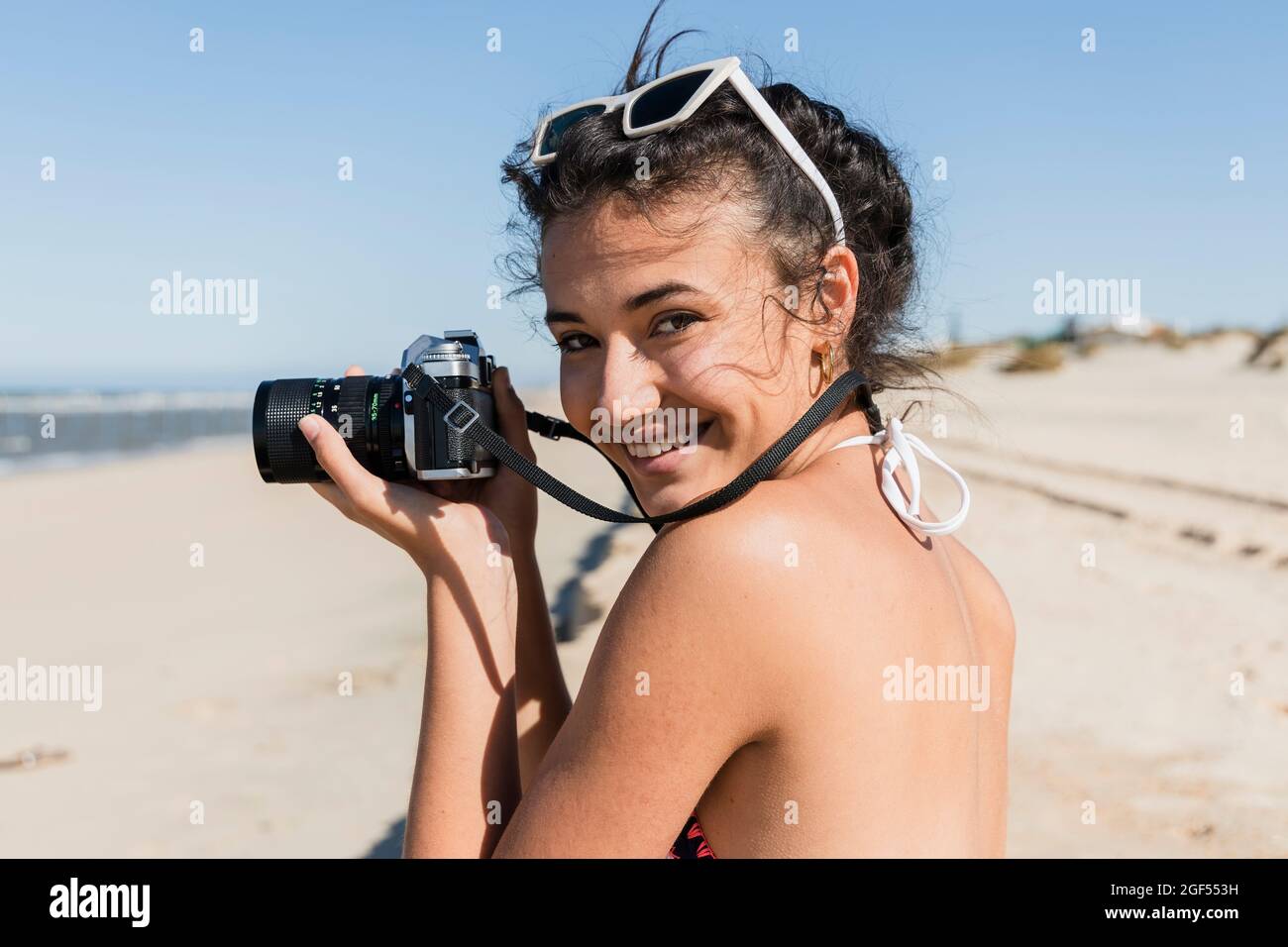 Jeune femme souriante avec appareil photo debout à la plage par beau temps Banque D'Images