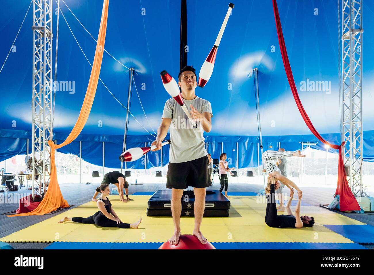 Épinglette de jonglage d'acrobat mâle tout en étant debout sur le ballon avec les athlètes pratiquant en arrière-plan au cirque Banque D'Images