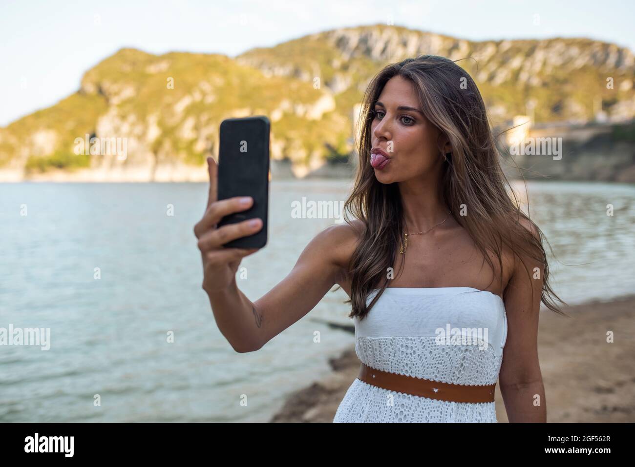 Jeune femme qui colle à la langue tout en prenant le selfie à travers le téléphone intelligent à Pantano de Santa Ana, Castillonroy, Espagne Banque D'Images