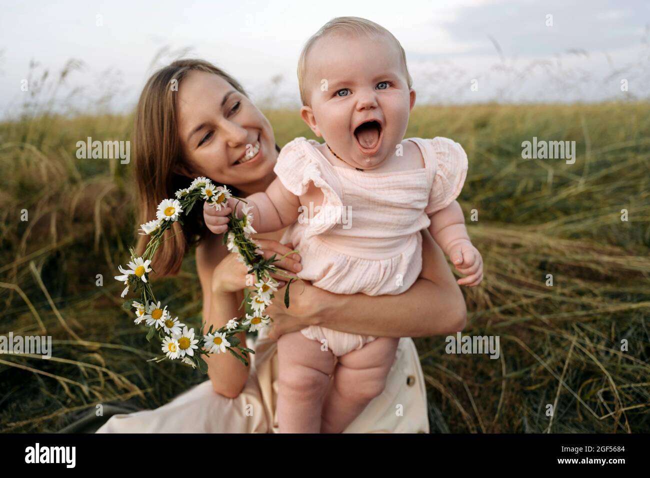 Mère souriante tenant bébé fille joyeuse avec tiara de fleur dans le champ Banque D'Images