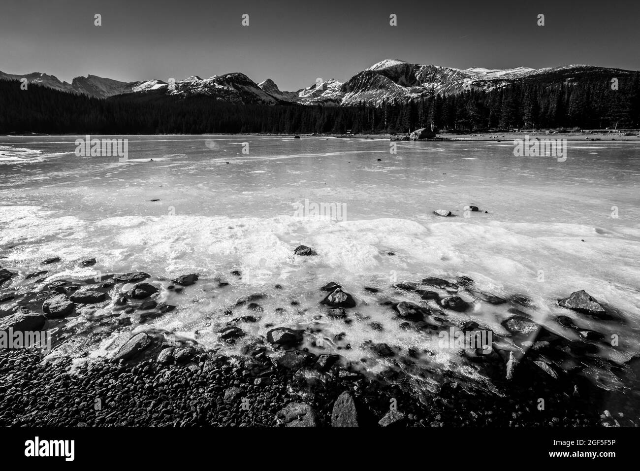Un paysage large et à faible angle de vue d'un lac gelé couvert de glace le long de la rive avec des pins et des montagnes en arrière-plan en noir et blanc Banque D'Images