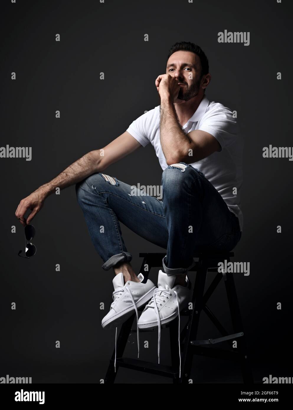 Homme assidu dans un polo blanc, jeans et baskets modernes et déchirés assis sur une chaise haute, escabeau Banque D'Images