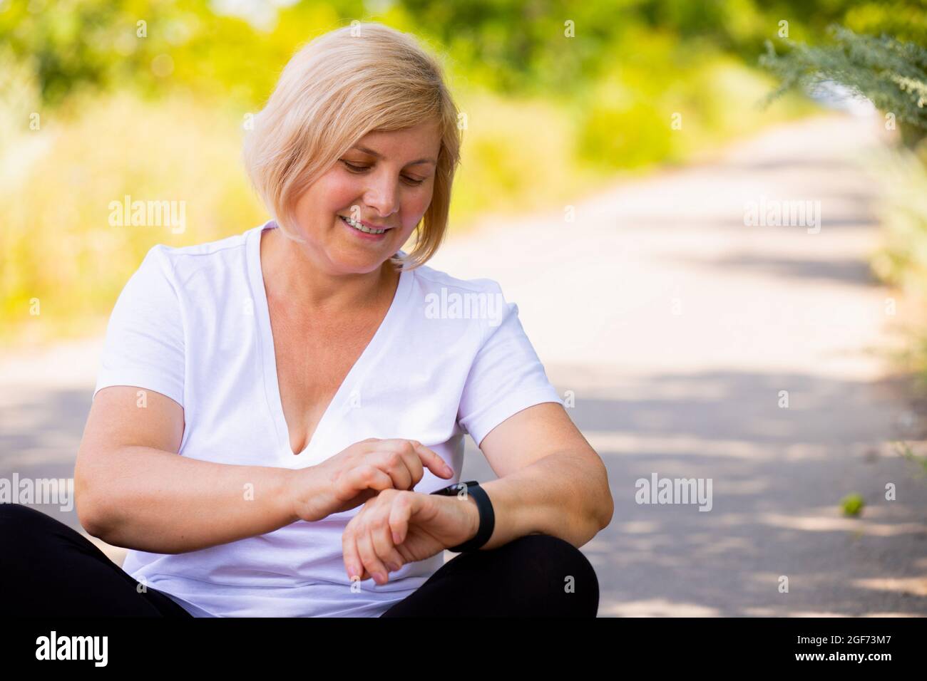 Gros plan d'une femme sportive assise avec des pieds croisés sur le chemin de la forêt, pointant sur sa montre numérique et souriant. Banque D'Images