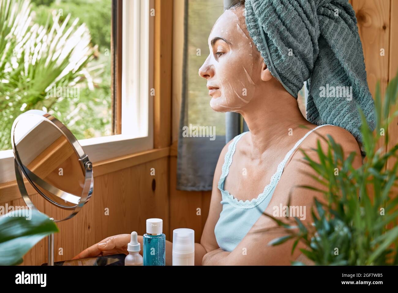 Soin de la peau. Femme d'âge moyen avec un masque hydratant en feuille dans son visage regarde à travers la fenêtre. Soulever le masque anti-rides sous les yeux. Collagène m Banque D'Images