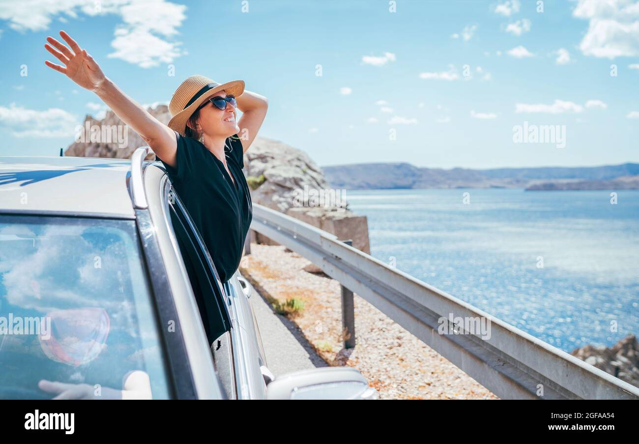 Portrait de femme joyeuse appréciant le voyage en bord de mer. Vêtue d'une robe noire, chapeau de paille et lunettes de soleil, elle ouvrit les bras et brillant avec le happine Banque D'Images