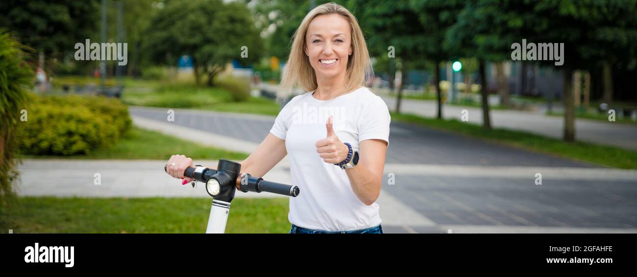 Femme avec un kick-scooter électrique debout dans la rue de la ville faisant un geste de pouce vers le haut. Banque D'Images