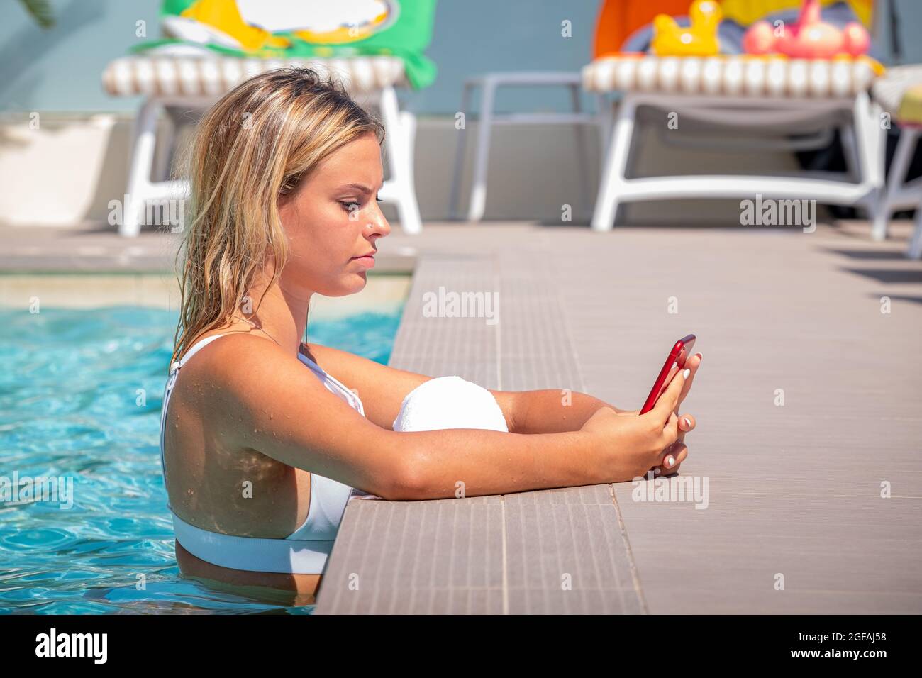 Bonne fille de caucase dans la piscine sourit tout en visiophonie et surfer sur Internet avec son smartphone rouge - Portrait bonne fille se relaxant dans la natation Banque D'Images