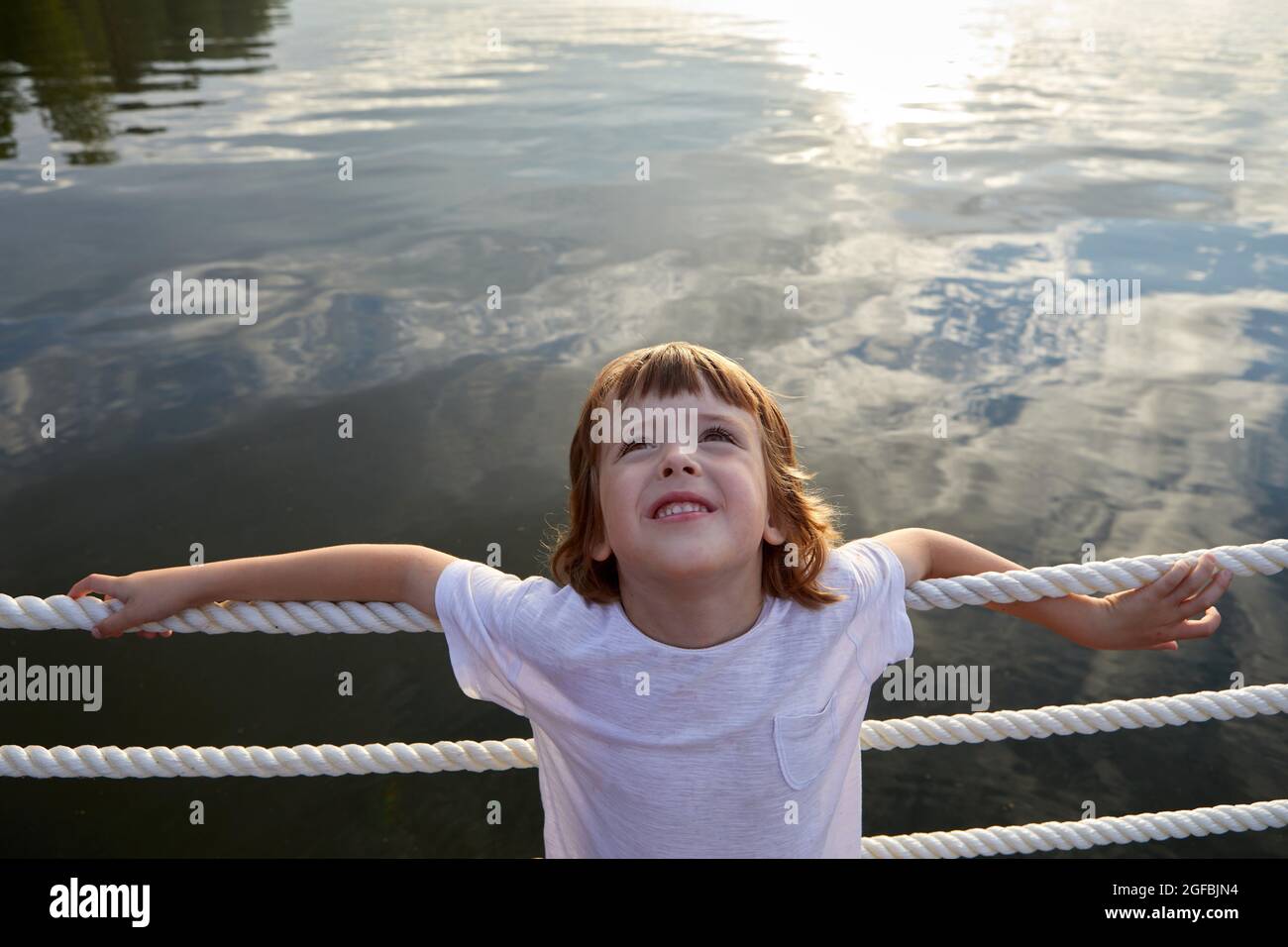 Grand angle de mignon garçon dans t-shirt blanc se pentant sur la barrière de corde et regardant vers le haut tout en se reposant près de l'eau propre de l'étang en été Banque D'Images