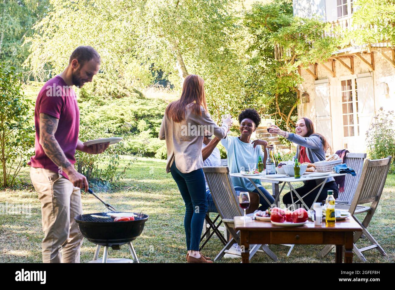 Jeune homme préparant de la nourriture au barbecue par ses amis assis à table Banque D'Images