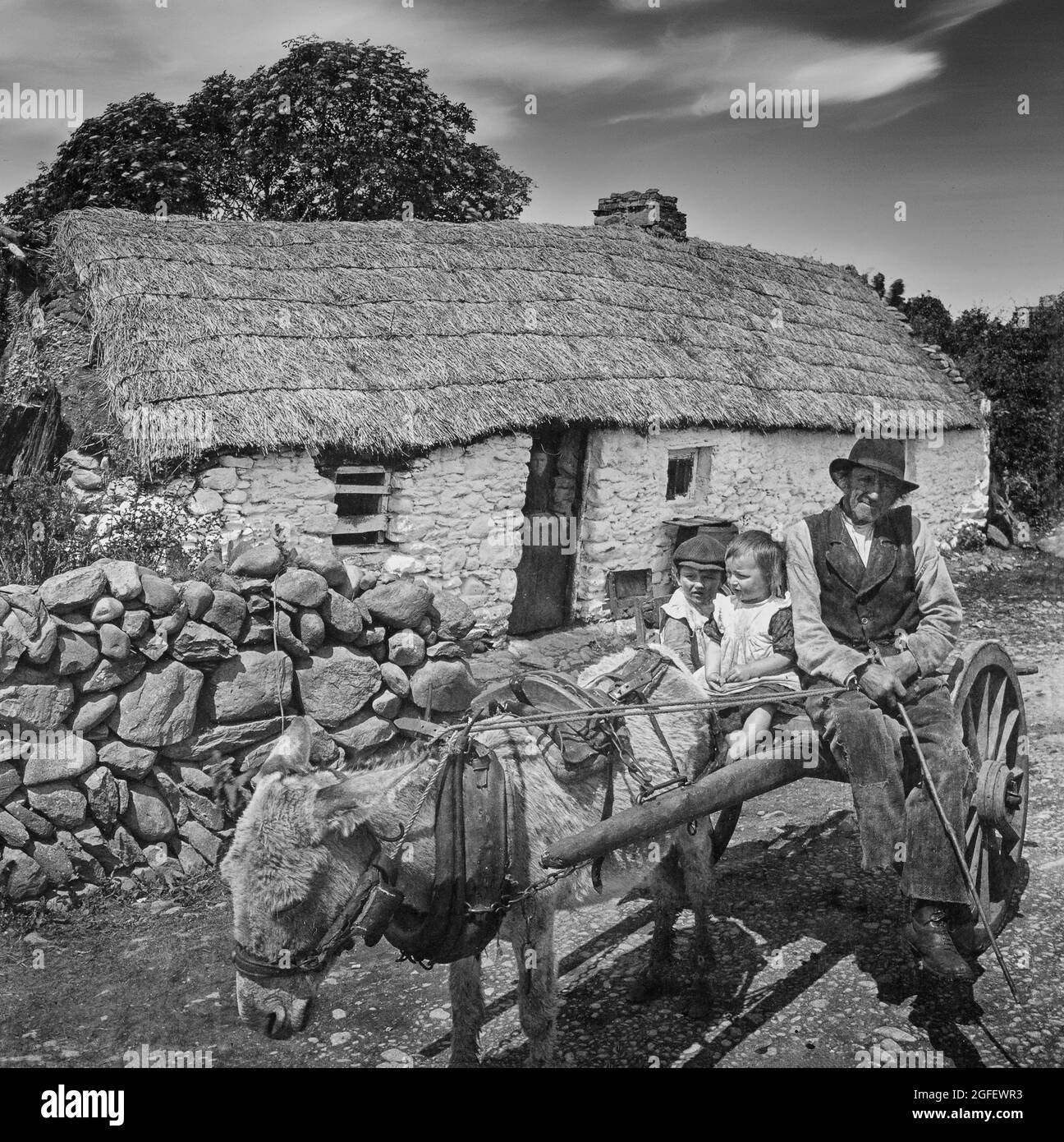 Vue du début du XXe siècle d'un fermier emportant des enfants sur une charrette à âne, tandis que sa femme regarde depuis la porte stable d'un traditionnel chaume sur la côte ouest de l'Irlande Banque D'Images