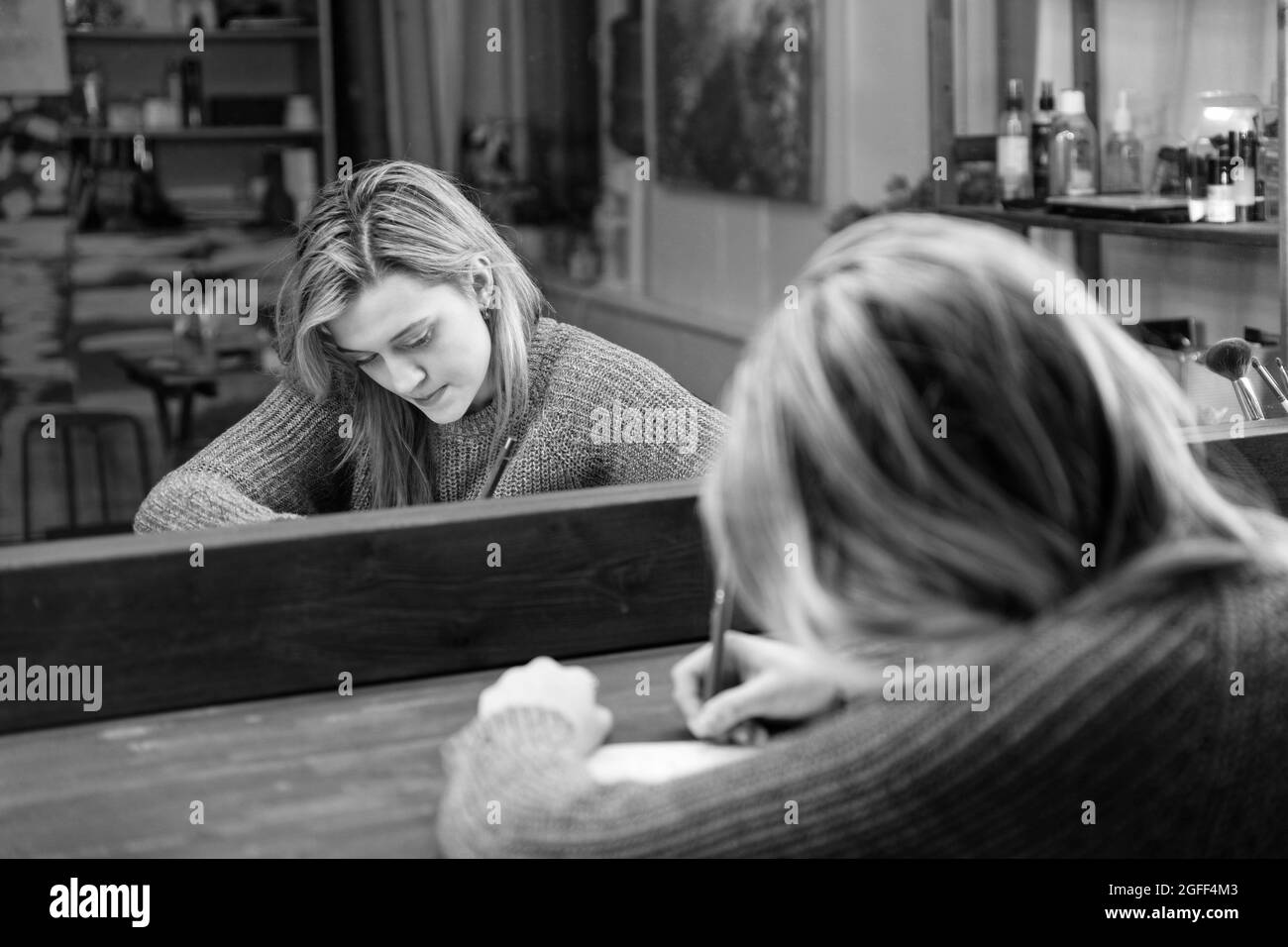 Une jeune femme s'assoit à une table devant un miroir et écrit quelque chose avec un stylo dans un bloc-notes. Mise au point peu profonde. Noir et blanc. Banque D'Images