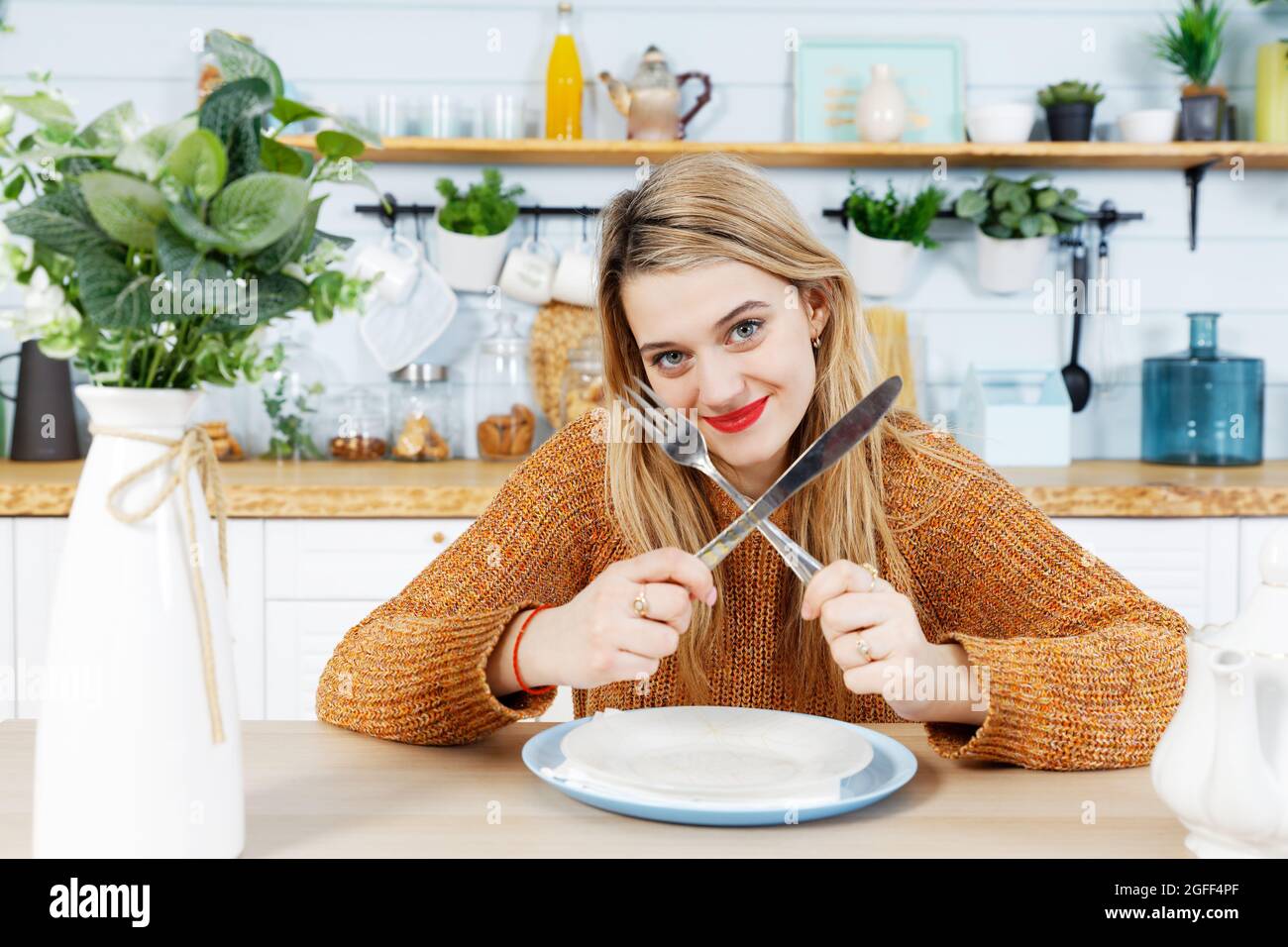 Jeune femme attrayante assise à une table dans la cuisine avec une assiette vide et tient une fourchette et un couteau les ont croisés entre eux Banque D'Images Jeune femme attrayante assise à une table dans la cuisine avec une assiette vide et tient une fourchette et un couteau les ont croisés entre eux Banque D'Images